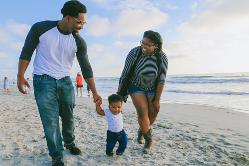 Imagen de un padre y una madre afroamericanos en la playa sosteniendo las manos de su hijo pequeño mientras caminan por la arena.